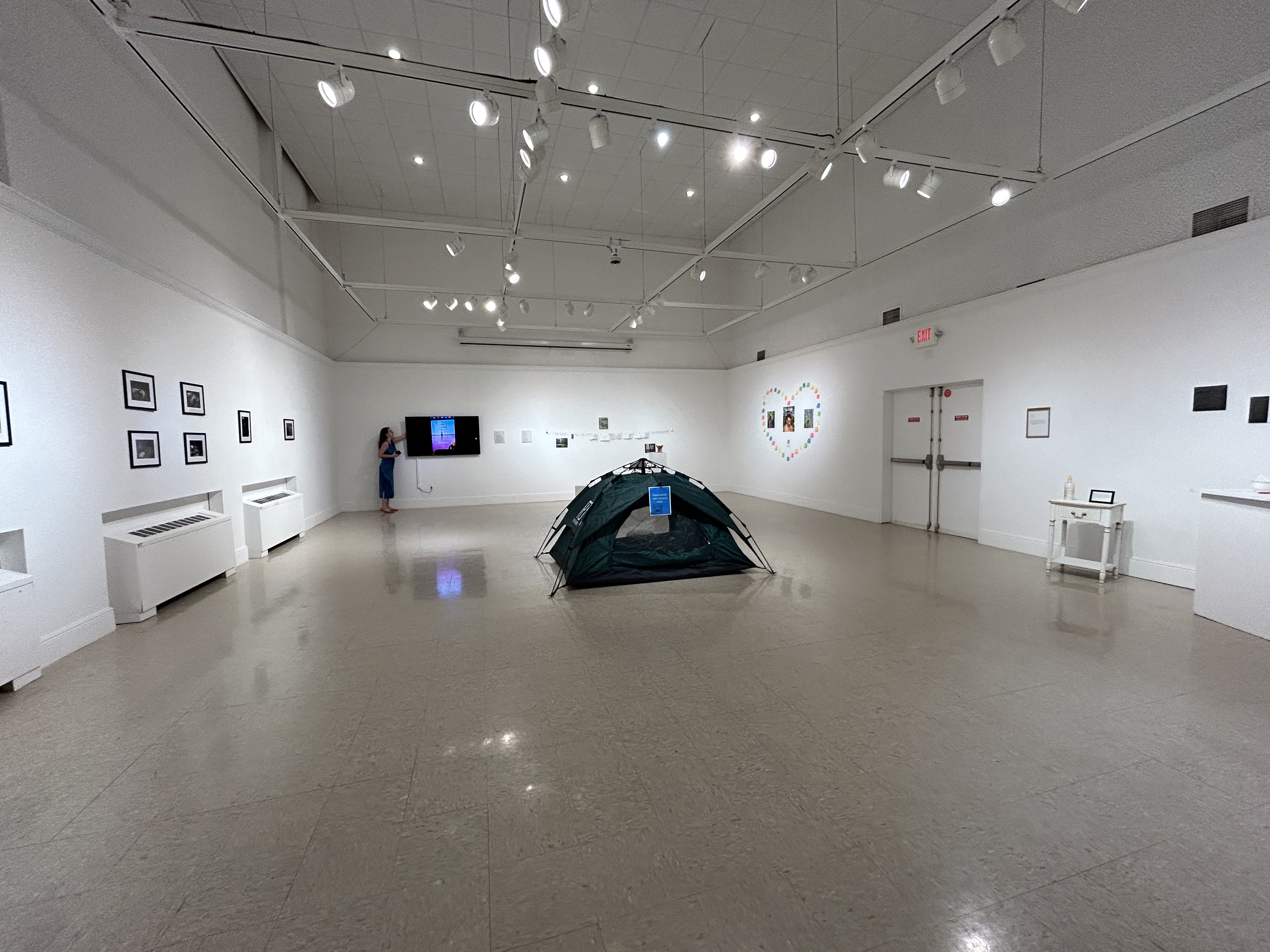 A gallery with white walls and cement floors hosts a tent at the center and various images along its walls.